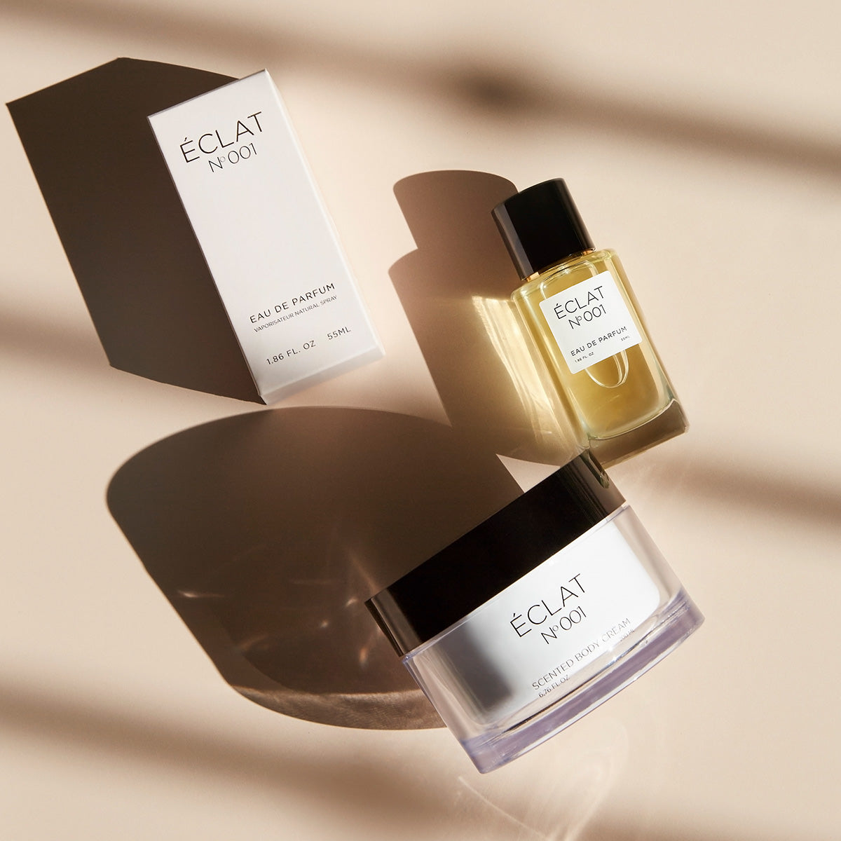 ÉCLAT No. 001 women’s fragrance set featuring a 55ml Eau de Parfum bottle, a matching light pink packaging box, and a scented body cream jar with black lid, all elegantly arranged on a beige surface with soft shadows.