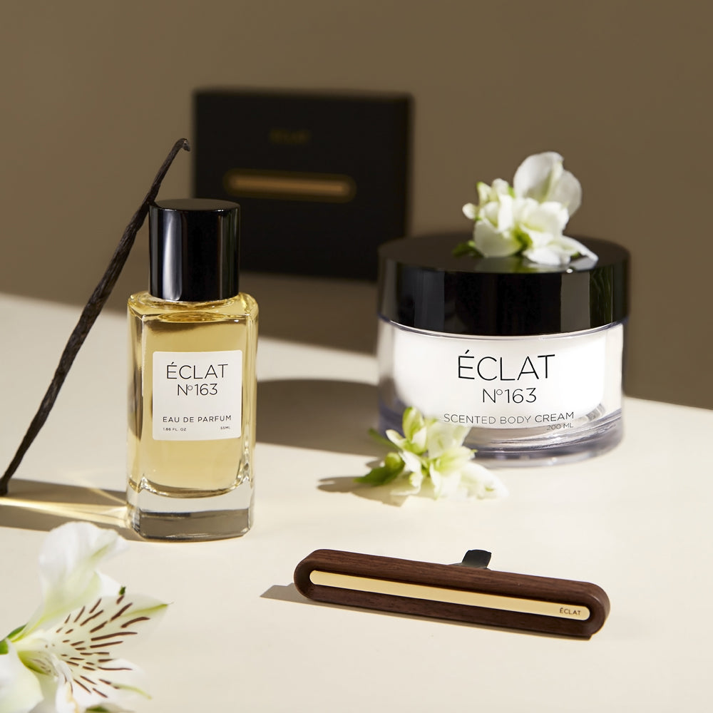 ÉCLAT Nr. 163 Damenduftkollektion, einschließlich Eau de Parfum, duftender Körpercreme und einem hölzernen Autoduftdiffusor, gestaltet mit weißen Blüten und Vanilleschote für eine sanfte, elegante Präsentation auf einer cremig-beigen Oberfläche.