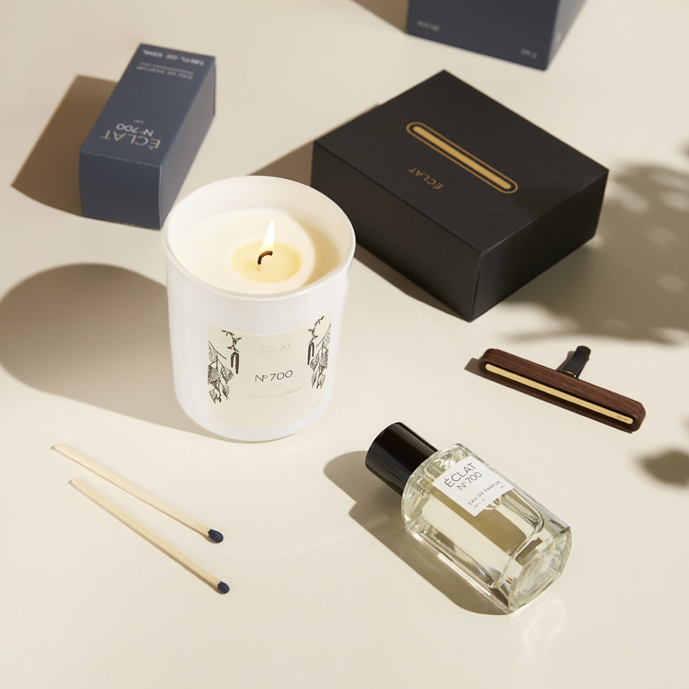 Flatlay of the ÉCLAT No. 700 fragrance set, featuring the Eau de Parfum, a lit scented candle, the matching dark blue perfume box, a car scent diffuser in dark wood with golden detail, and its packaging. Two matches lie beside the candle, all arranged on a softly lit cream surface.