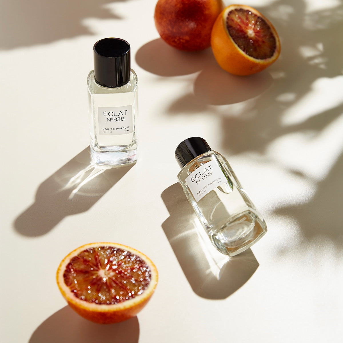 Two flacons of ÉCLAT No. 938 unisex perfume styled on a light surface with fresh blood oranges. The minimalist glass bottles with black caps reflect the vibrant essence of the fragrance.