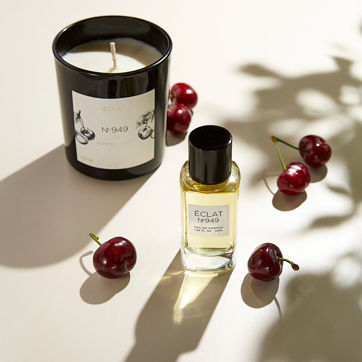 ÉCLAT No. 949 perfume and scented candle set arranged with fresh cherries on a bright surface. The black glass candle features a cherry illustration, while the matching perfume bottle has a yellow-toned fragrance, a black cap, and a white label.