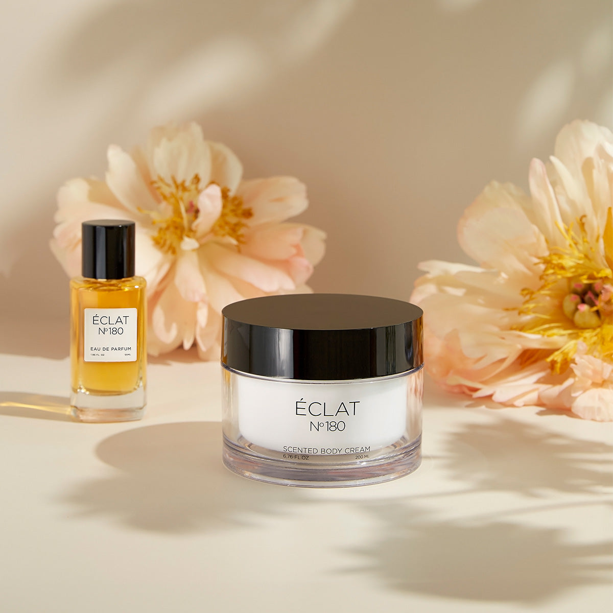ÉCLAT Orchid & Patchouli Set mit einem klaren Glasparfümflakon und einem Cremetiegel mit schwarzem Deckel, umgeben von zarten Blumen.
