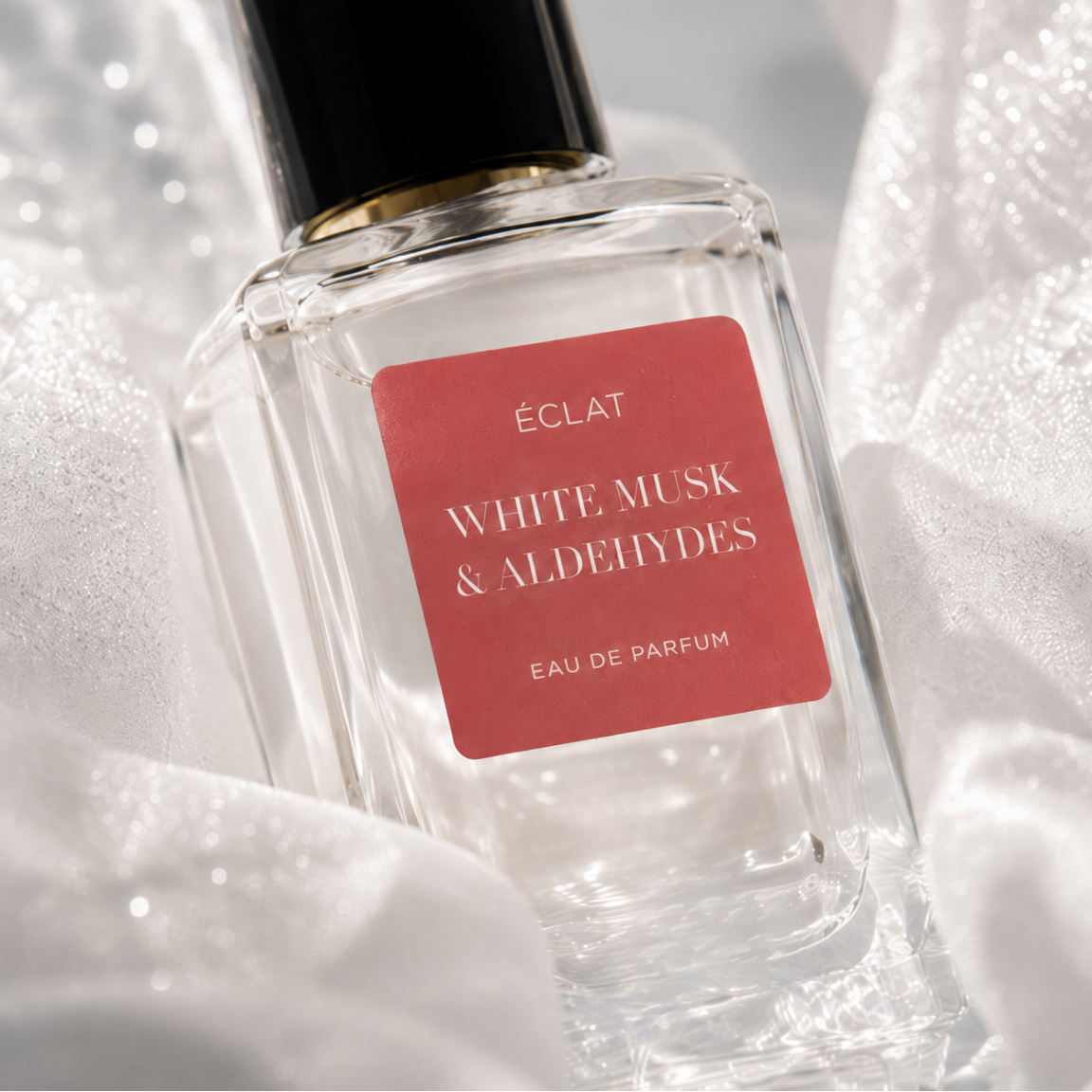 Ein facettierter Glasflakon von ÉCLAT White Musk & Aldehydes mit rotem Etikett und schwarzer Kappe, umgeben von zartem weißen Stoff.
