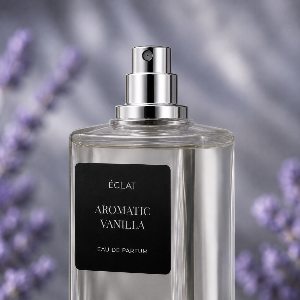Ein klarer Glasflakon von ÉCLAT Aromatic Vanilla mit polierter silberner Sprühkappe und mattschwarzem Etikett vor verschwommenem Lavendel-Hintergrund.