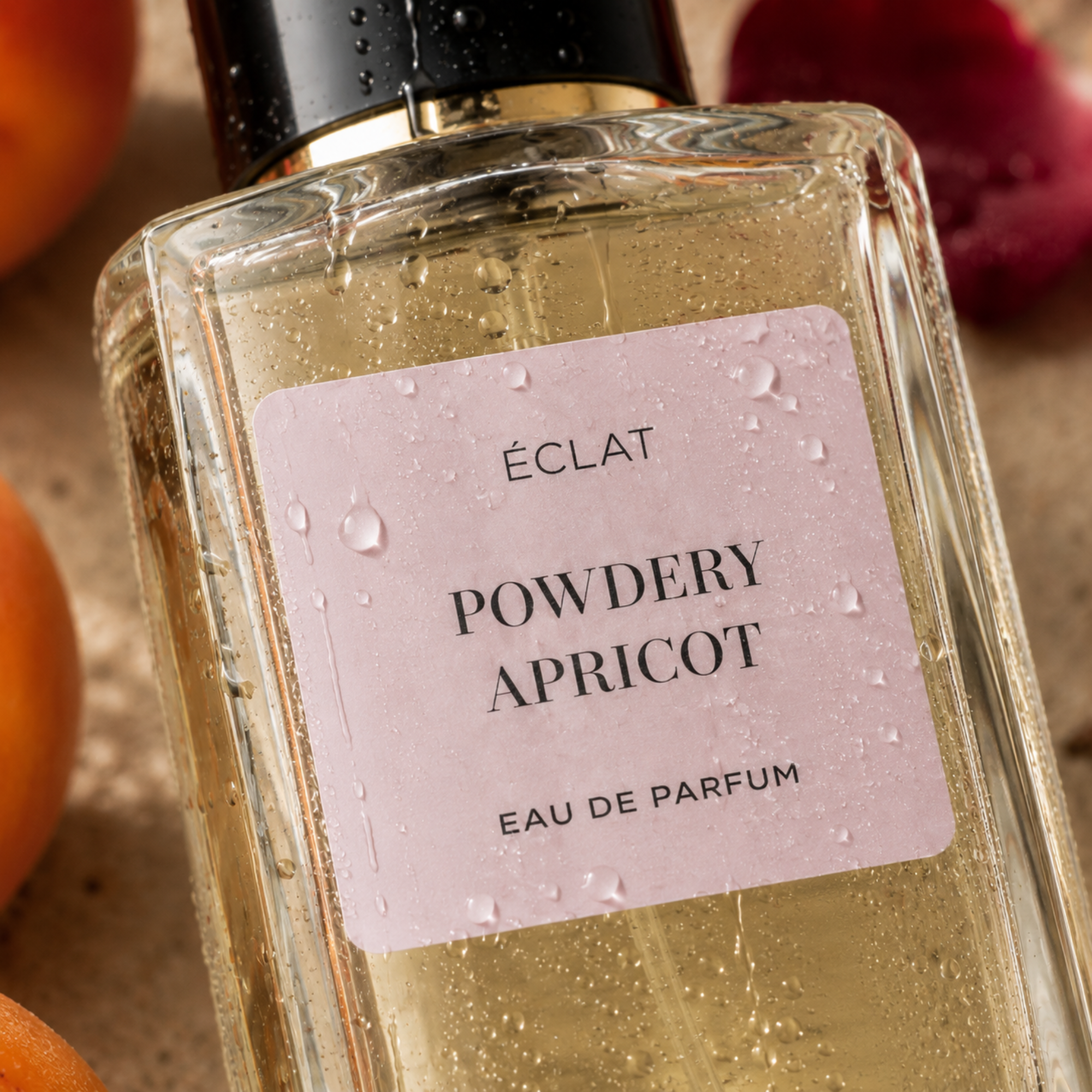 Facettierter Glasflakon von ÉCLAT Powdery Apricot mit hellrosa Etikett und schwarzer Kappe, umgeben von Aprikose und roten Blütenblättern.