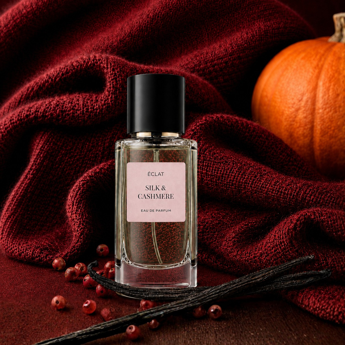 Ein facettierter Glasflakon von ÉCLAT Silk & Cashmere mit schwarzer Kappe, arrangiert vor tiefrotem Stoff, umgeben von Vanilleschoten, Beeren und einem Kürbis.
