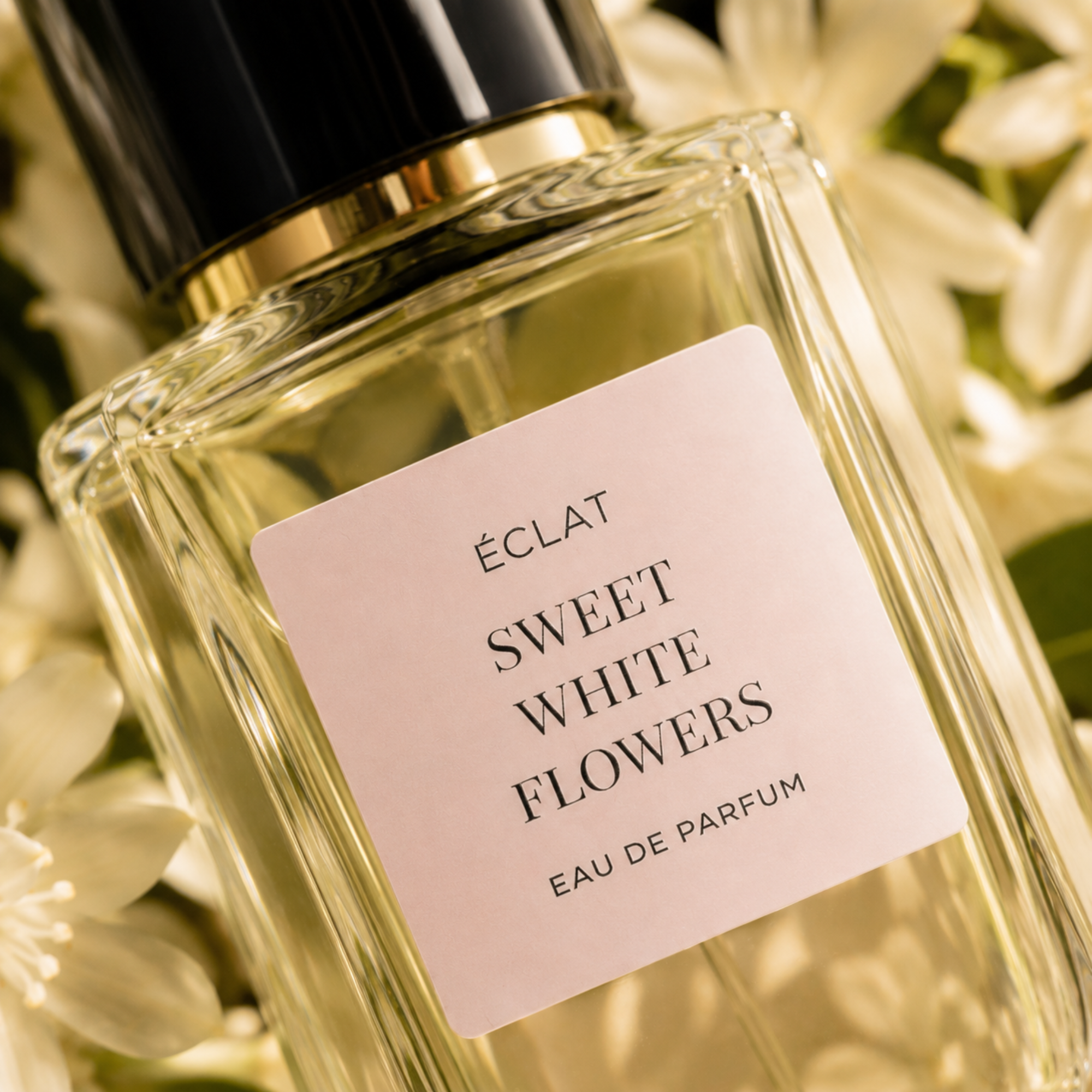 Ein facettierter Glasflakon von ÉCLAT Sweet White Flowers mit einem sanft rosa Etikett und einer schwarz-goldenen Kappe, umgeben von weißen Blüten.