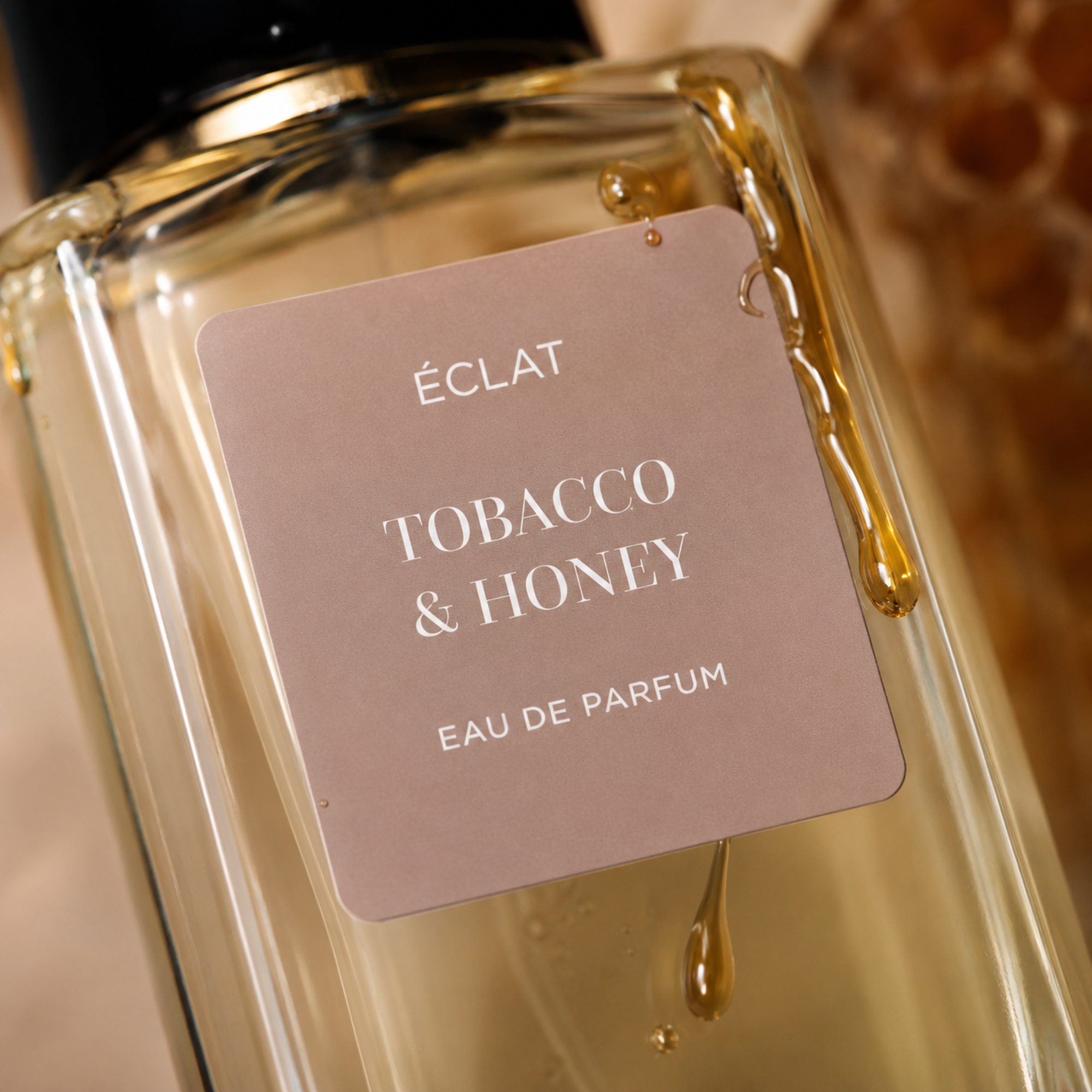 Parfümflakon ÉCLAT Tobacco & Honey mit hellbraunem mattem Etikett und einem Honigtropfen, der über die Glasfläche fließt.