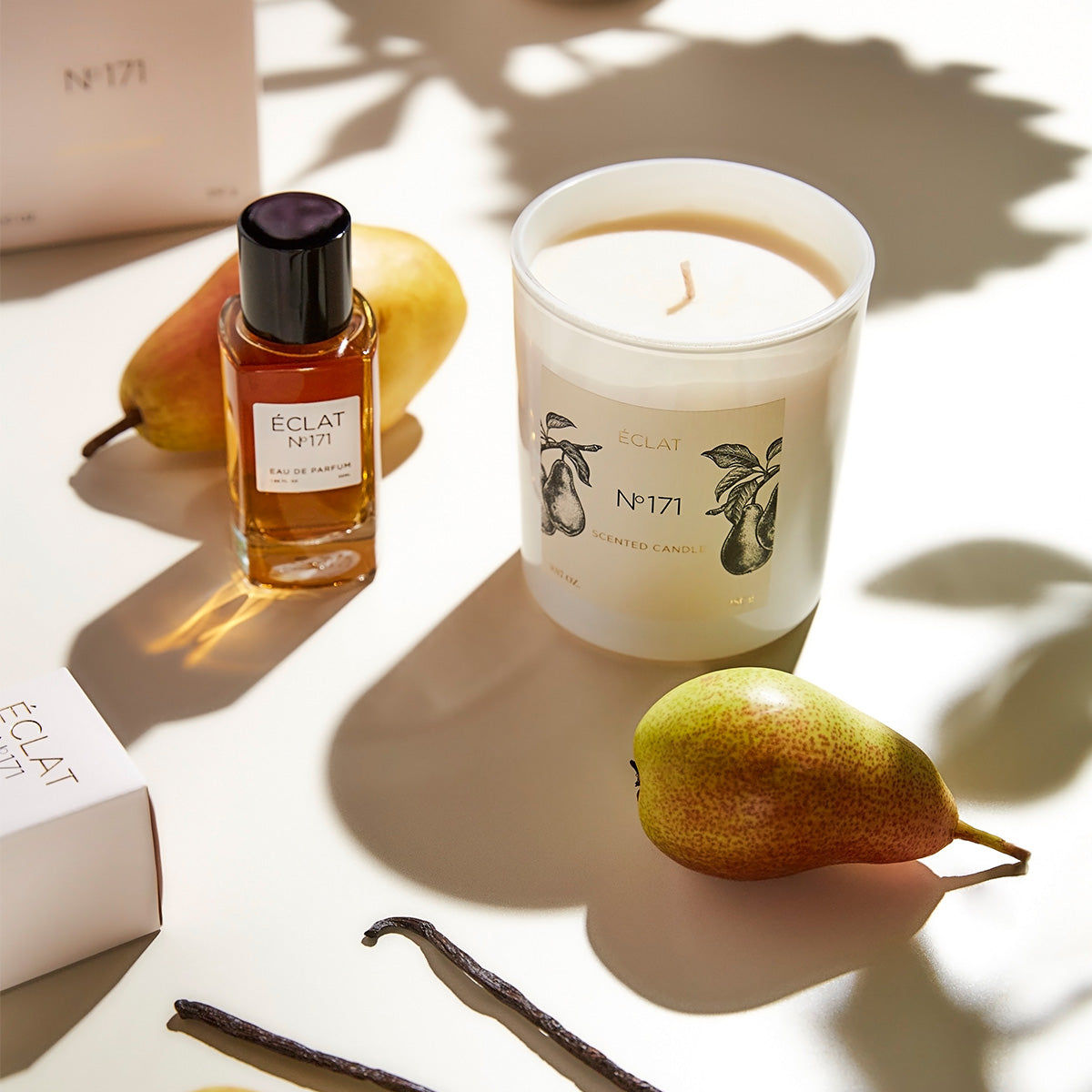 Glasflakon des ÉCLAT Blackcurrant & Praline Parfums mit schwarzer Kappe neben einer eleganten weißen Kerze und Birne auf hellem Untergrund.