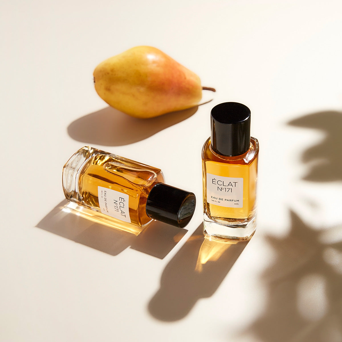 ÉCLAT Blackcurrant & Praline in eleganten Glasflakons mit schwarzen Verschlüssen, neben einer reifen Birne, weiche Schatten werfend.
