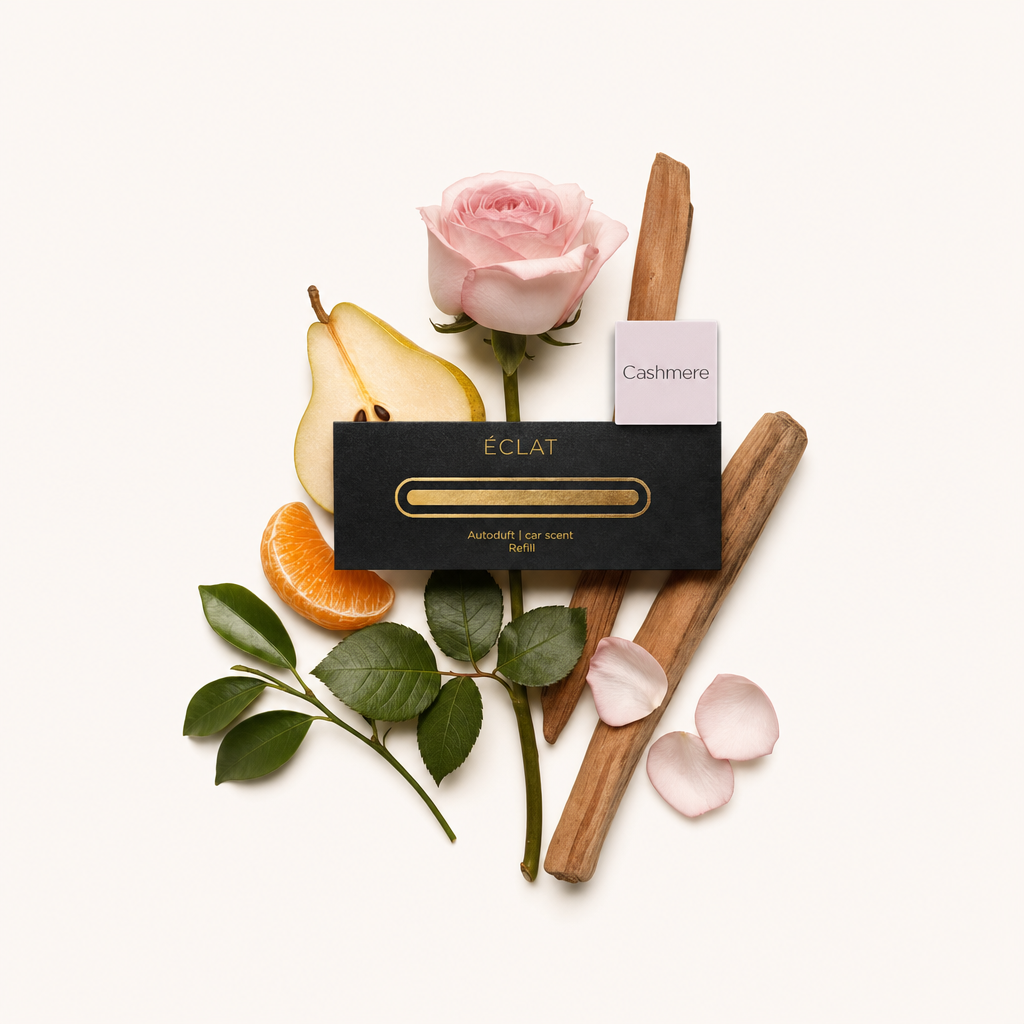 Ein zartes Arrangement aus einer rosafarbenen Rose, einer aufgeschnittenen Birne, einem Mandarinenstück, zwei Holzstäben, grünen Blättern und verstreuten Blütenblättern, neben einer schwarzen Kappe von ÉCLAT mit einem 'Cashmere'-Etikett.