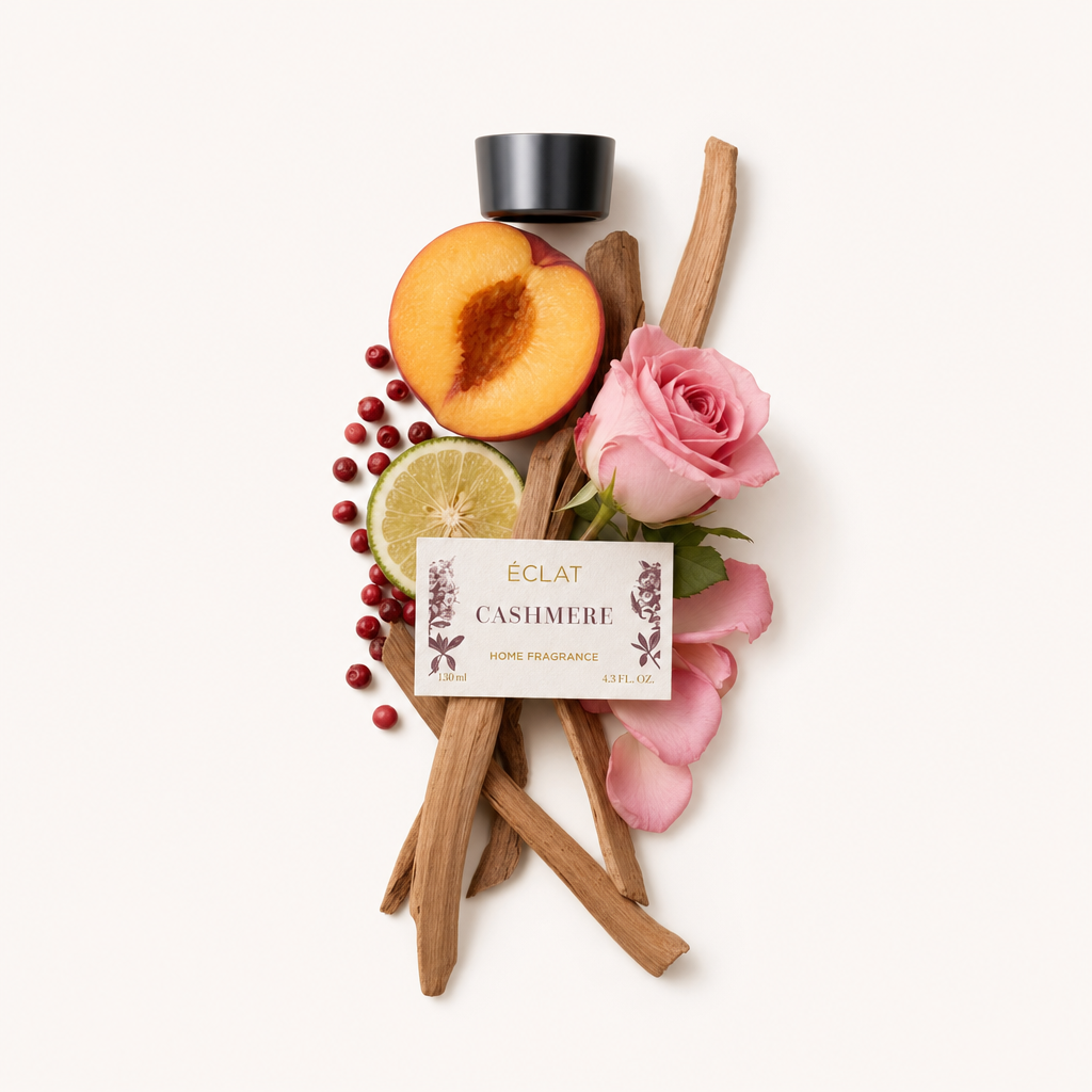 Ein Arrangement aus Sandelholzstäben, einer halben Pfirsich, einer Limettenscheibe, rosa Rose und roten Beeren umgibt eine schwarze Kappe mit der Aufschrift ÉCLAT Cashmere Room Fragrance Sticks.
