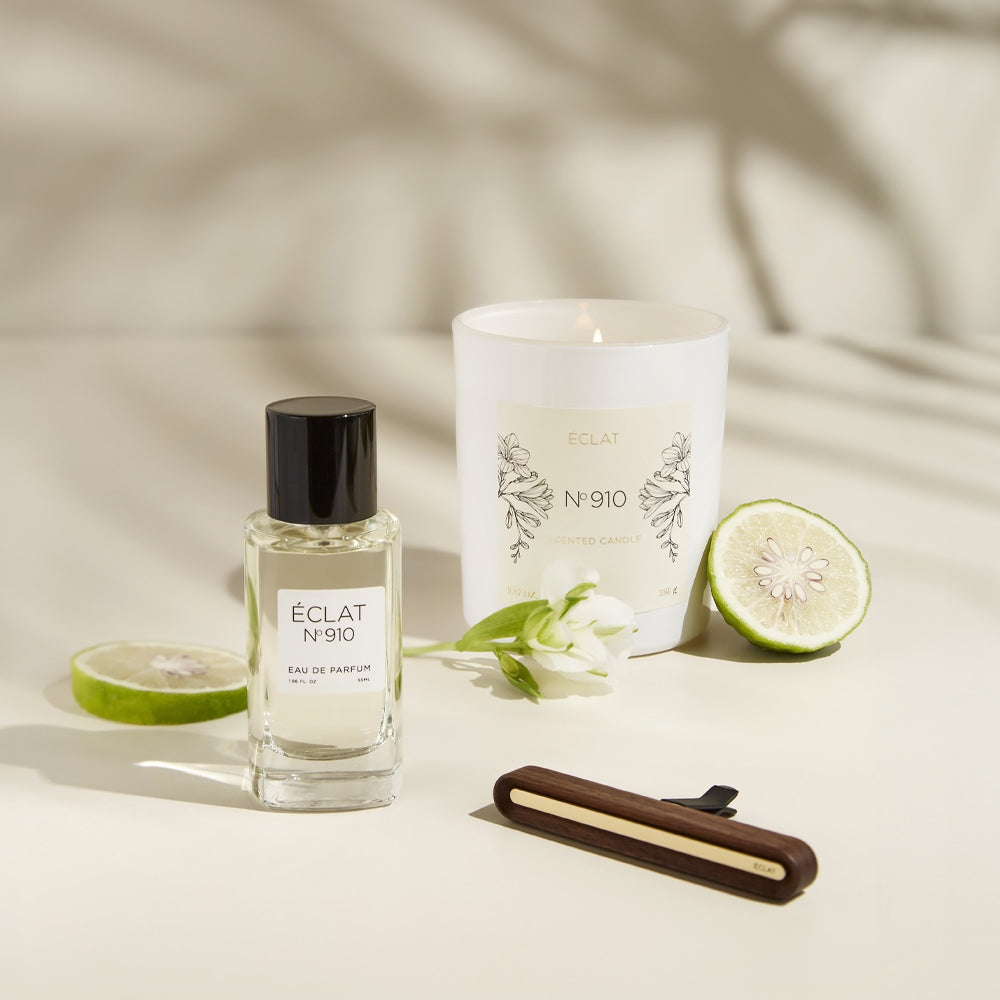 Ein facettierter Glasflakon des ÉCLAT Creamy White Parfums mit schwarzem Verschluss, neben einer weißen Duftkerze, Zitrusscheibe und Blume.