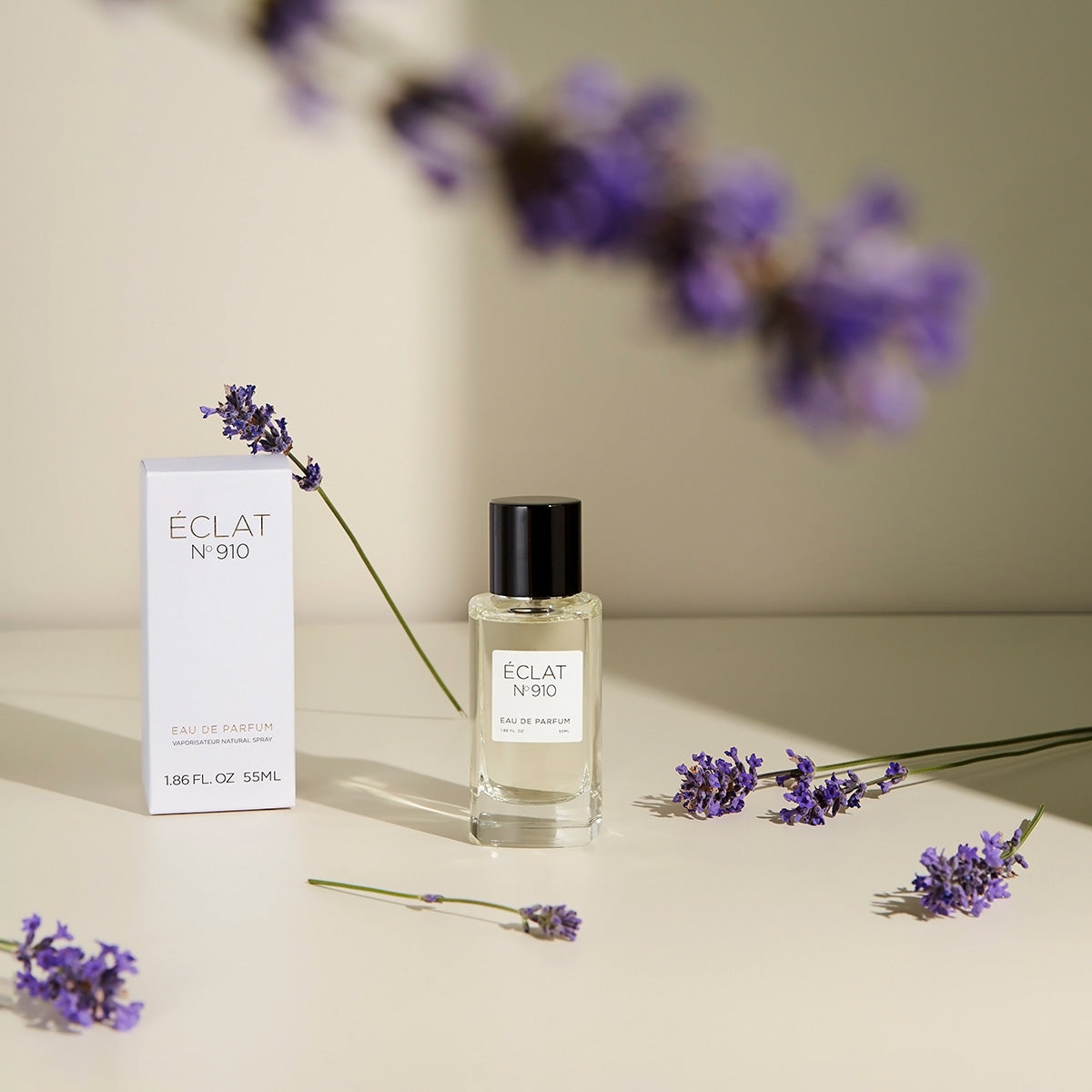 ÉCLAT Creamy White Parfum in einem klaren Glasflakon mit schwarzem Verschluss, umgeben von Lavendelzweigen, steht neben seiner eleganten weißen Box.