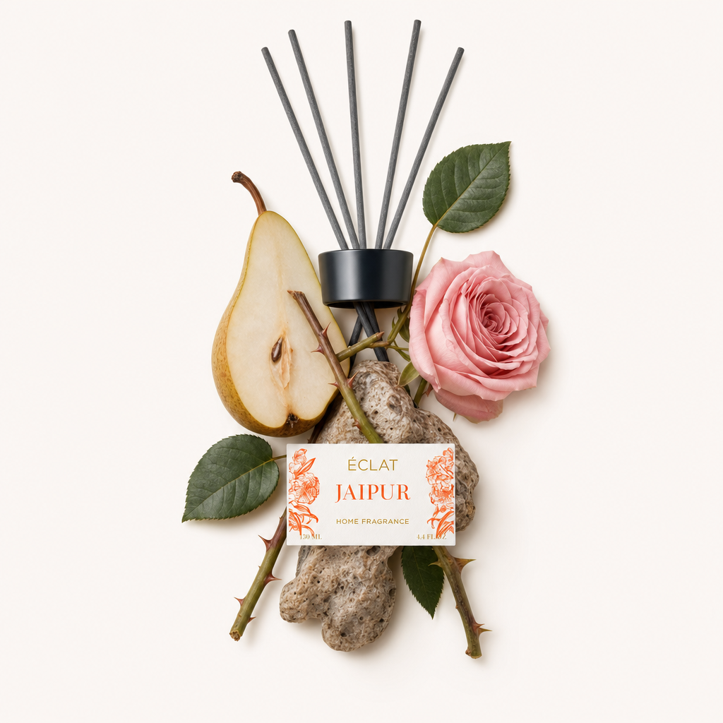 Ein Arrangement aus ÉCLAT Jaipur Duftnoten mit einer Rose, einer Birnenscheibe, grünen Blättern und strukturierten Steinen, neben einer schwarzen Kappe mit Etikett.