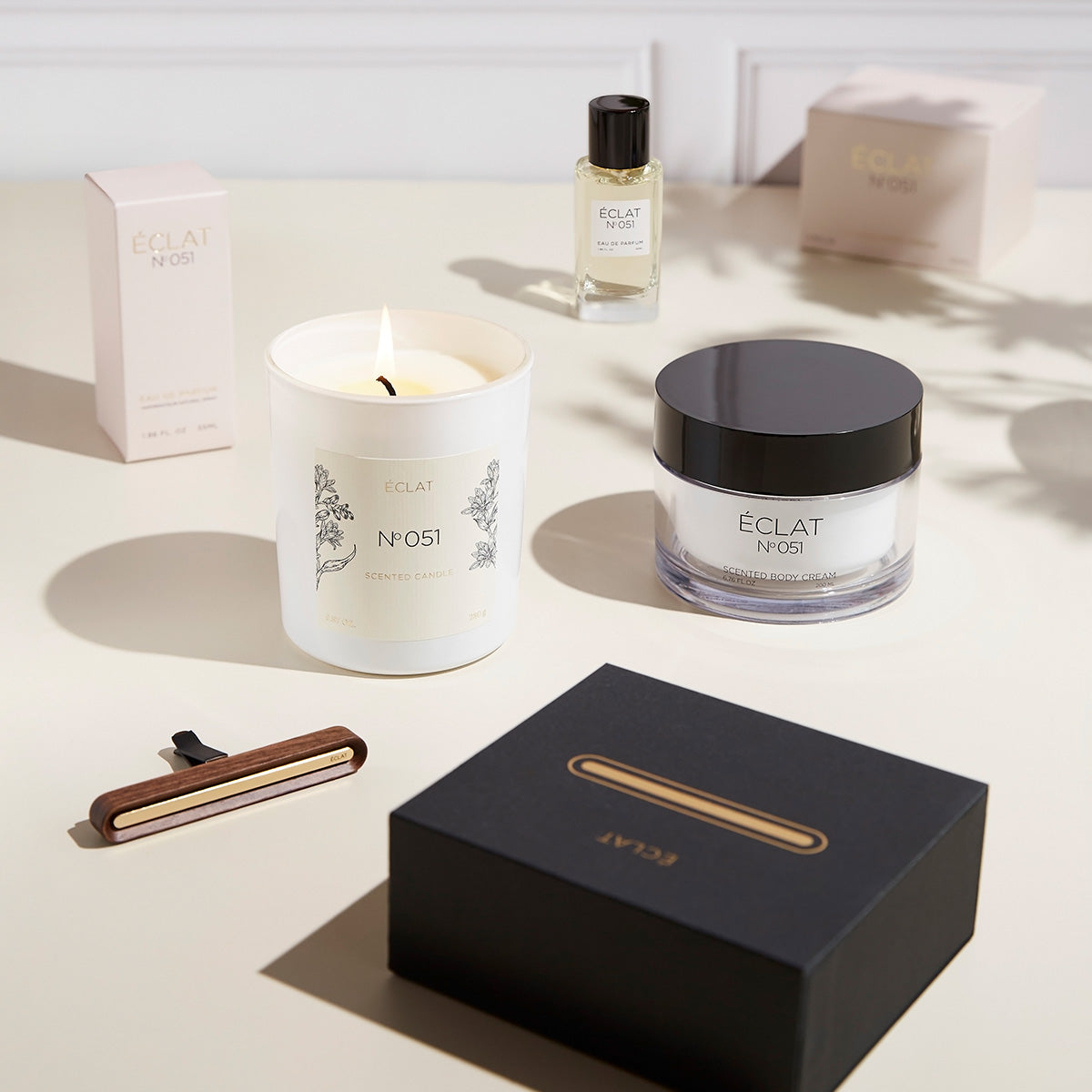 Eine elegante Komposition mit einer brennenden Duftkerze, einem facettierten Glastiegel mit dunklem Deckel, einem Parfümflakon und einer edlen Box, die ÉCLAT Oriental Almond verkörpert.