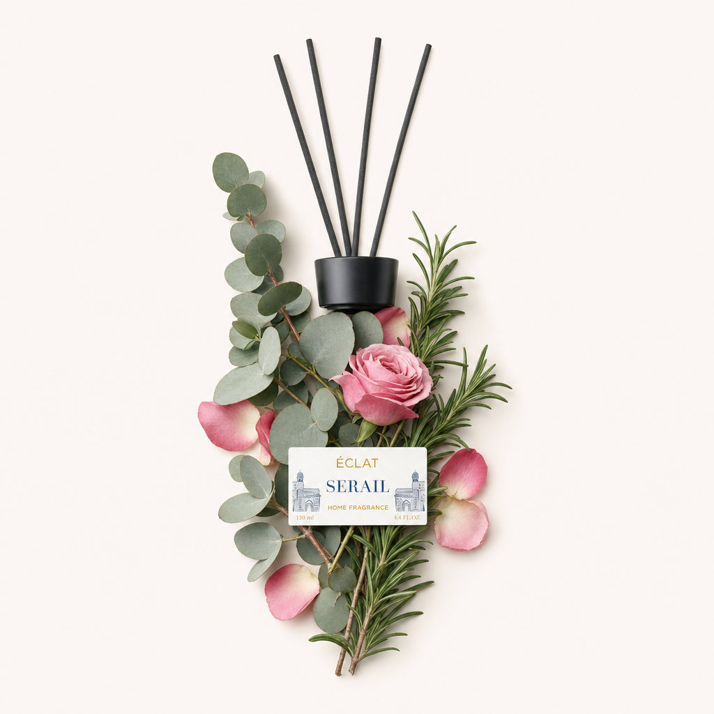 Ein Arrangement aus Eukalyptusblättern, Rosmarinzweigen, rosafarbenen Rosenblütenblättern und einer blühenden Rose umrahmt eine schwarze Kappe mit einem Etikett für ÉCLAT Serail Room Fragrance Sticks.