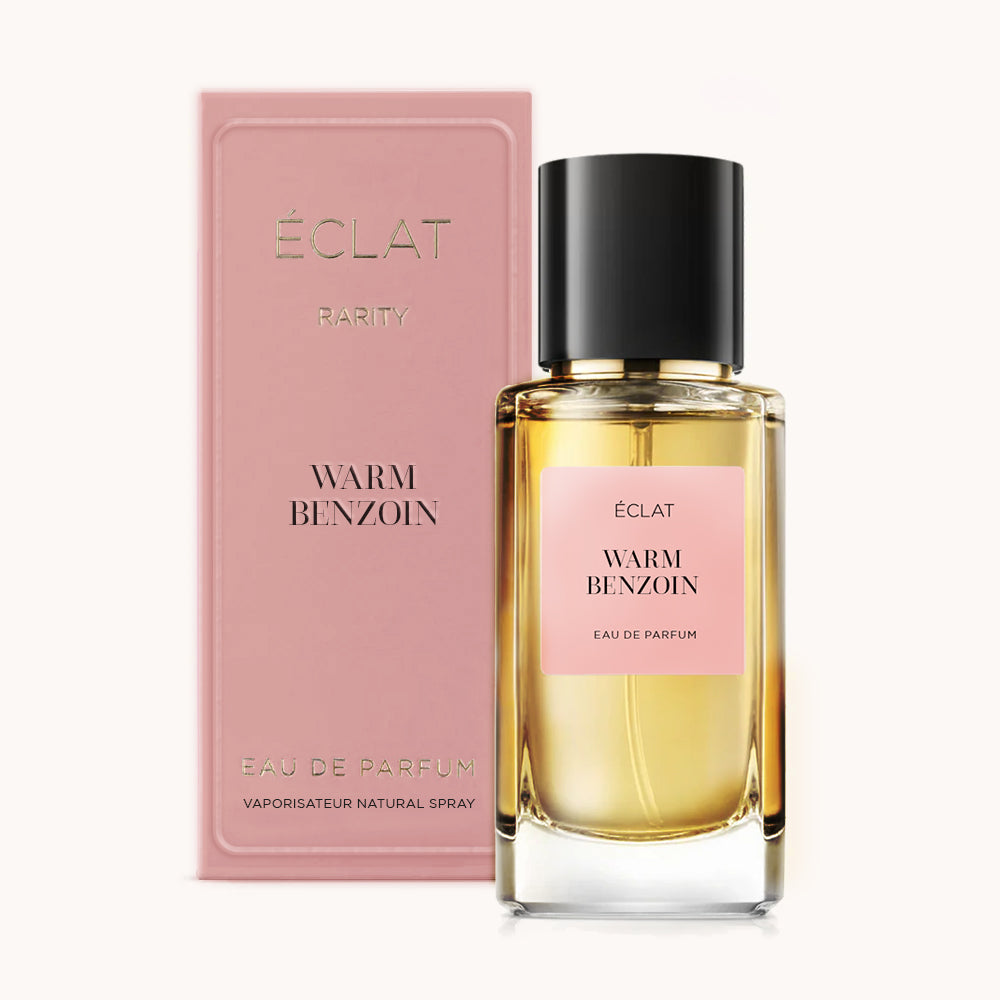 Das ÉCLAT Warm Benzoin Parfum wird mit einem transparenten Glasflakon mit schwarzem Verschluss und rosa Etikett gezeigt; daneben steht eine passende rosa Box.